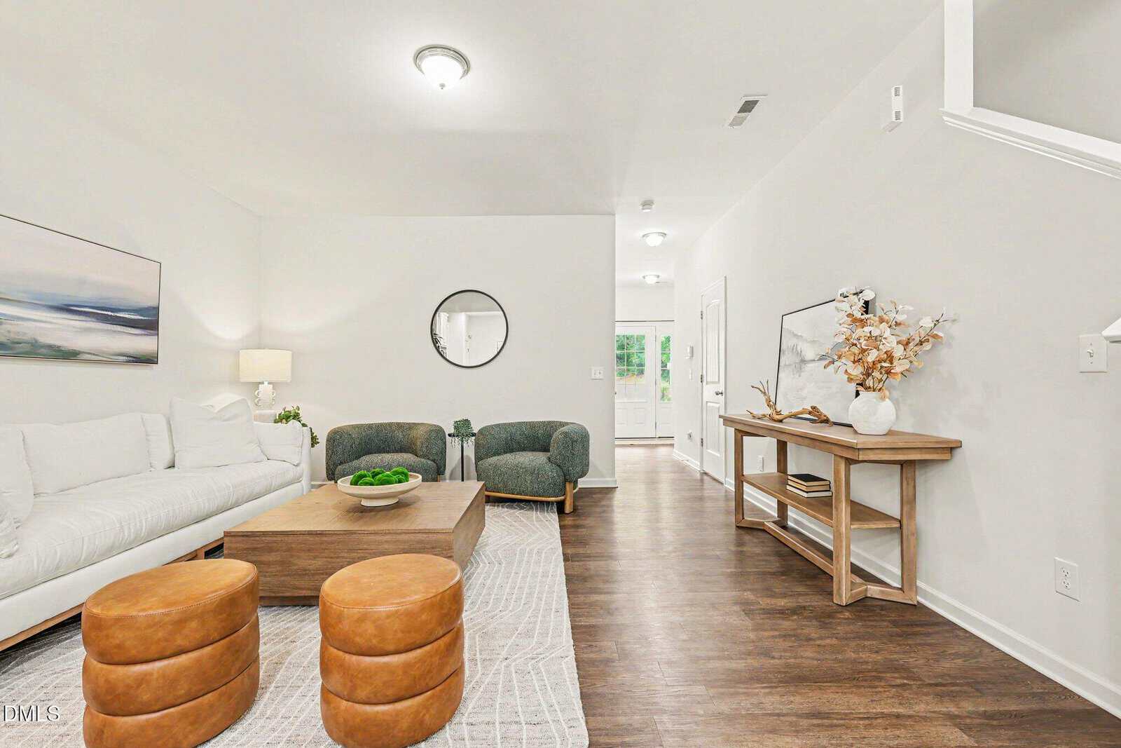 Modern living room with a white sofa, two green chairs, two round tan ottomans, a wooden coffee table, and sideboard with decor. Neutral colors and wooden floor with a light rug.