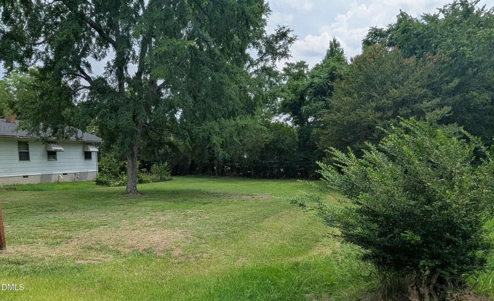 A grassy yard with scattered trees and bushes, next to a white house with a gray roof; the background features dense greenery and a partly cloudy sky.