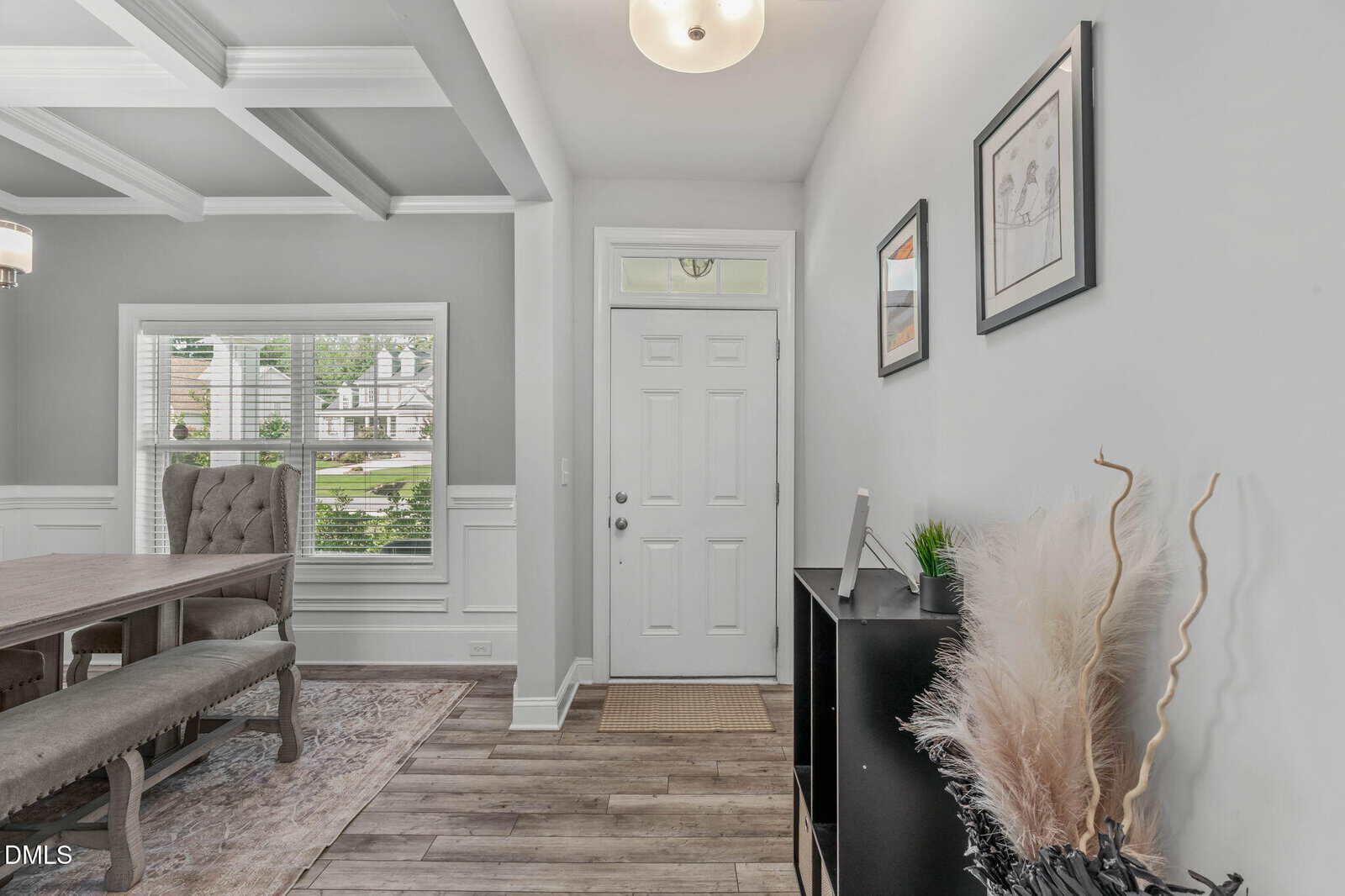 A bright entryway with a white front door, two framed pictures on a pale wall, a black shelf with decor, and a dining area with a table and bench by a large window with blinds.