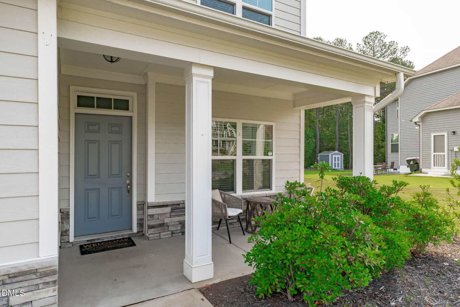 A house with a covered front porch, a gray door, white columns, two chairs with a small table, and green shrubs in the yard. Another house and a shed are visible in the background.