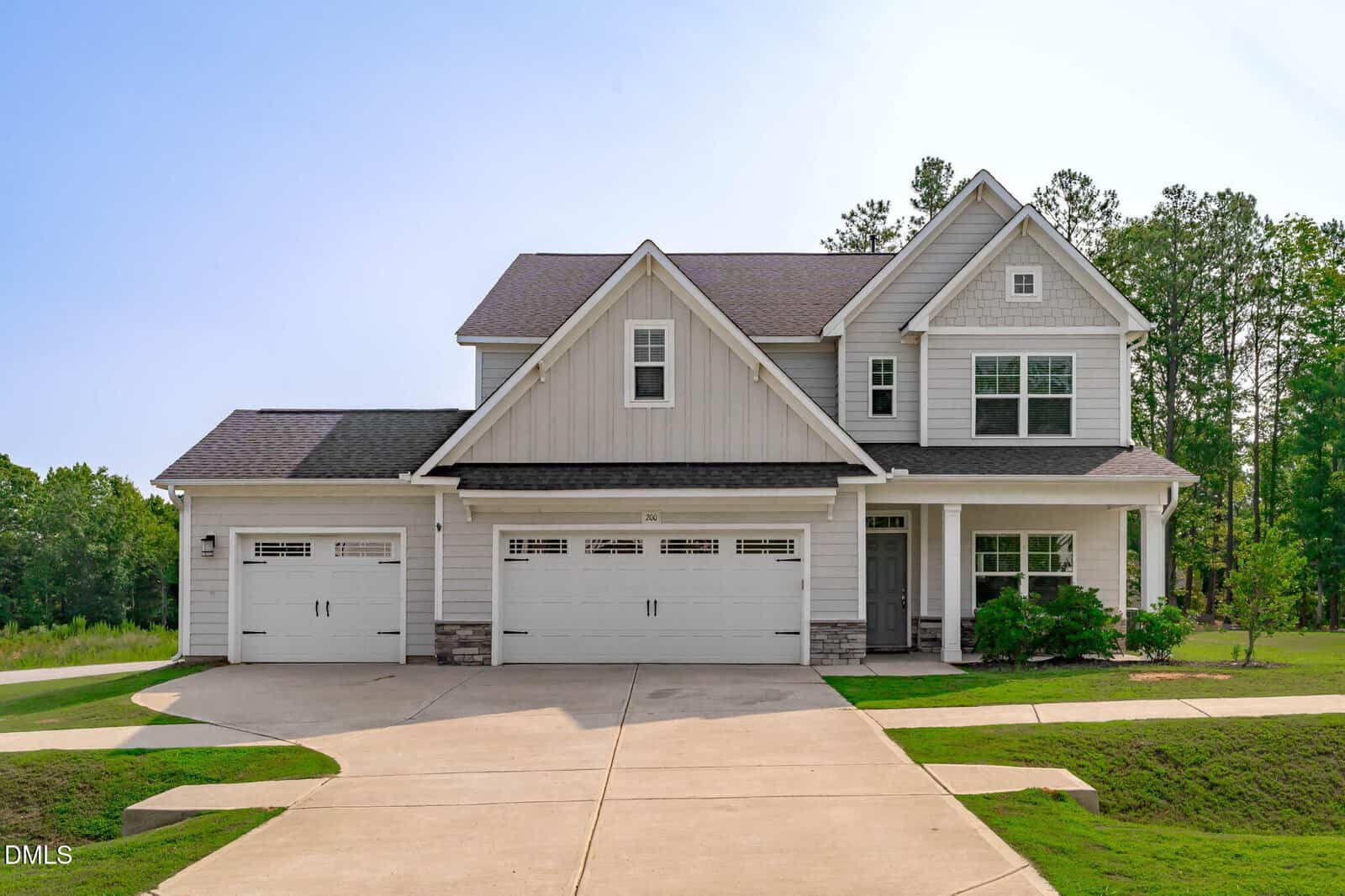 Two-story suburban house with light gray siding, three white garage doors, a covered front porch, and a large driveway. Located at 200 Sutherland Drive in Franklinton, this home is surrounded by green grass and trees under a clear sky.