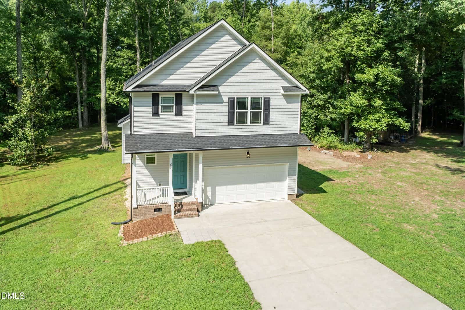 This two-story gray house with white trim at 202 Cardinal Court, Oxford, features an attached two-car garage, inviting front porch, and concrete driveway. Surrounded by a lush green lawn and mature trees in a peaceful wooded area, it offers a charming retreat on a sunny day.