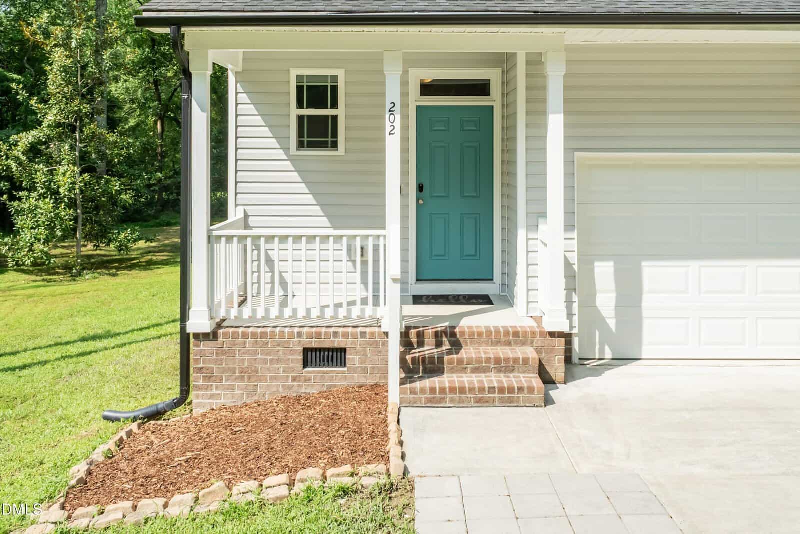 A small porch with white railings and steps leads to a blue front door on a light gray house with brick accents. The house has a garage, and a concrete path and grass yard are visible.