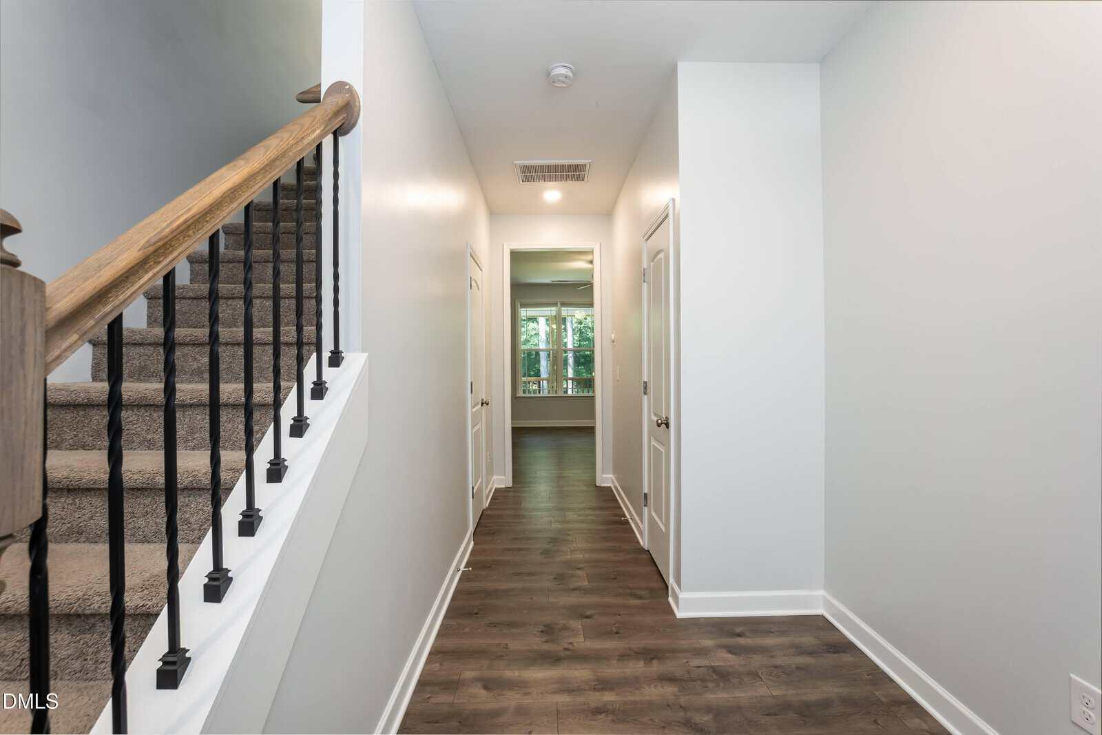 A hallway with wood flooring, white walls, a staircase with dark handrails on the left, and multiple doors leading to a bright room with large windows at the end.