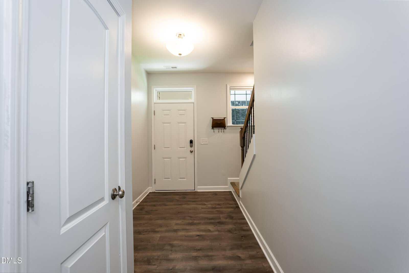 A hallway with wood flooring leads to a white front door. There is a staircase with a dark handrail on the right and windows above the stairs. White walls and a ceiling light create a bright, welcoming entryway.