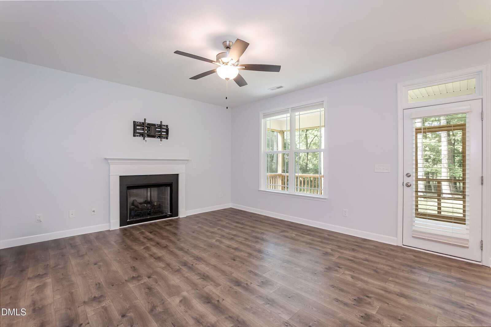 Bright living room with wood flooring, ceiling fan light, fireplace with mantel, wall mount, large window, and glass door leading to a screened porch with trees visible outside. Room is unfurnished and has light-colored walls.