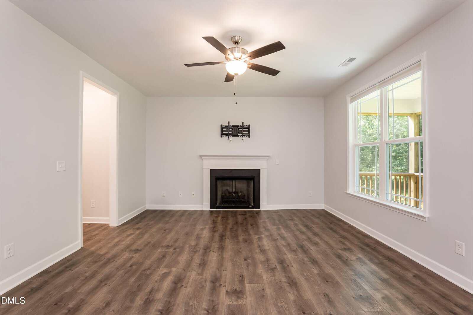 Bright, empty living room with wood floors, white walls, a ceiling fan, fireplace, and a large window showing a porch and greenery outside.