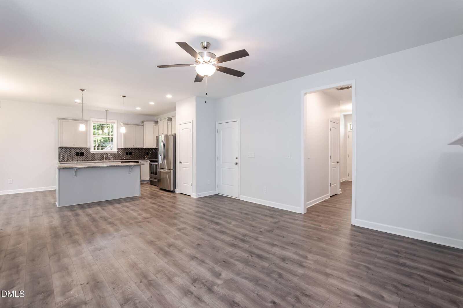 Open concept living area with gray wood floors, ceiling fan, and a modern kitchen featuring a large island, stainless steel appliances, white cabinets, and a tile backsplash. Hallway and multiple doors are visible.
