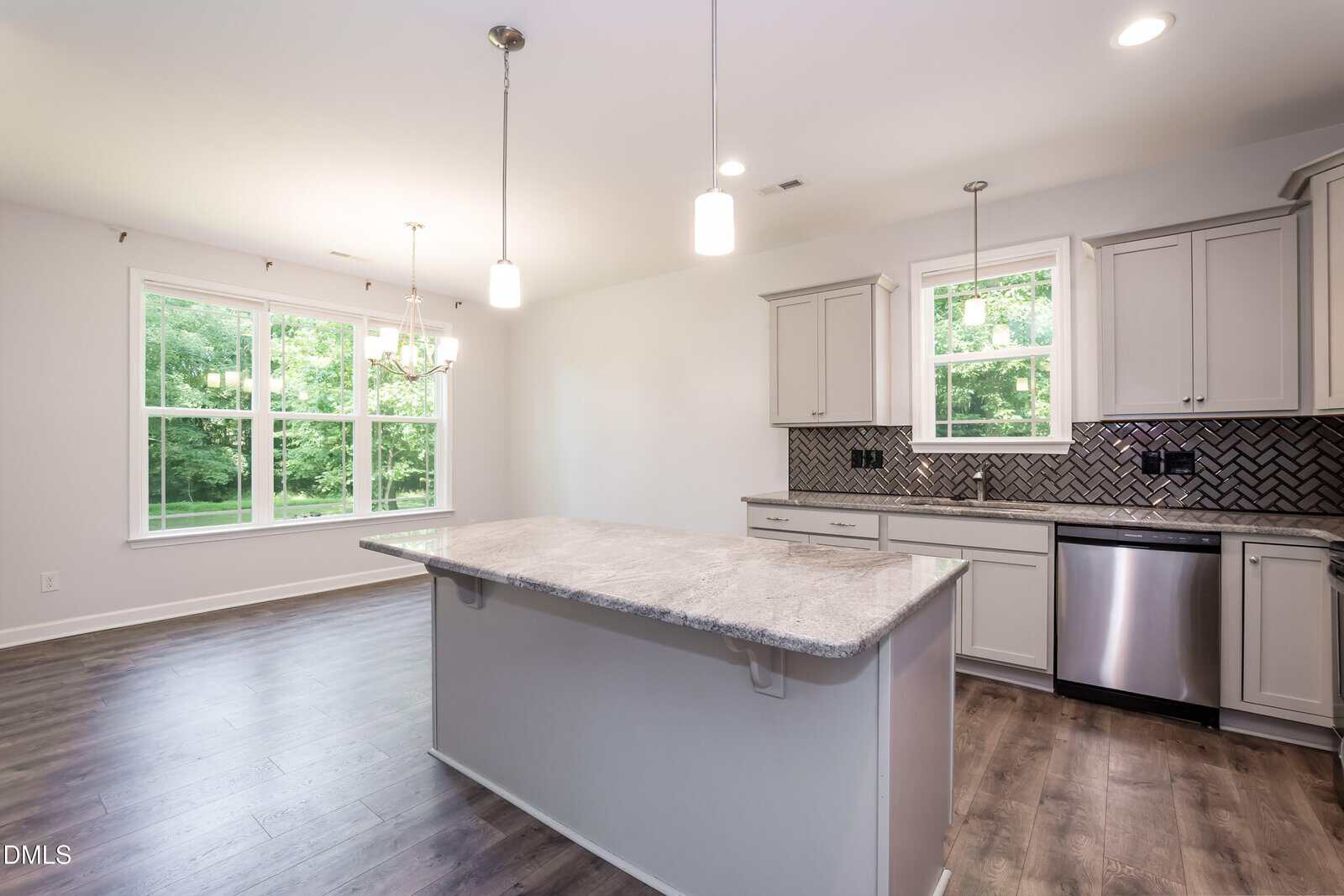 Bright kitchen with light gray cabinets, marble island, stainless steel appliances, herringbone tile backsplash, pendant lights, and large windows overlooking green trees. Dining area visible to the left.