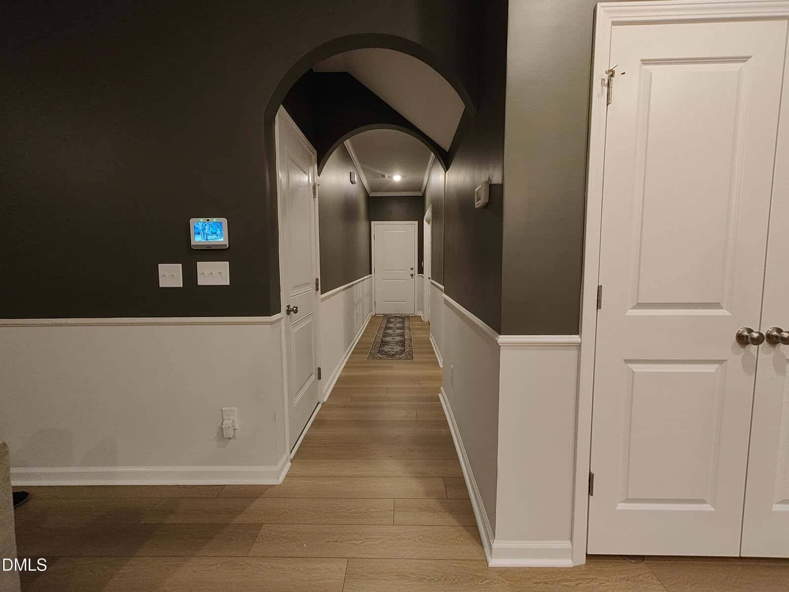 A hallway with arched ceilings, dark upper walls, white lower walls, light wood floors, white doors, a rug, and a wall-mounted thermostat.