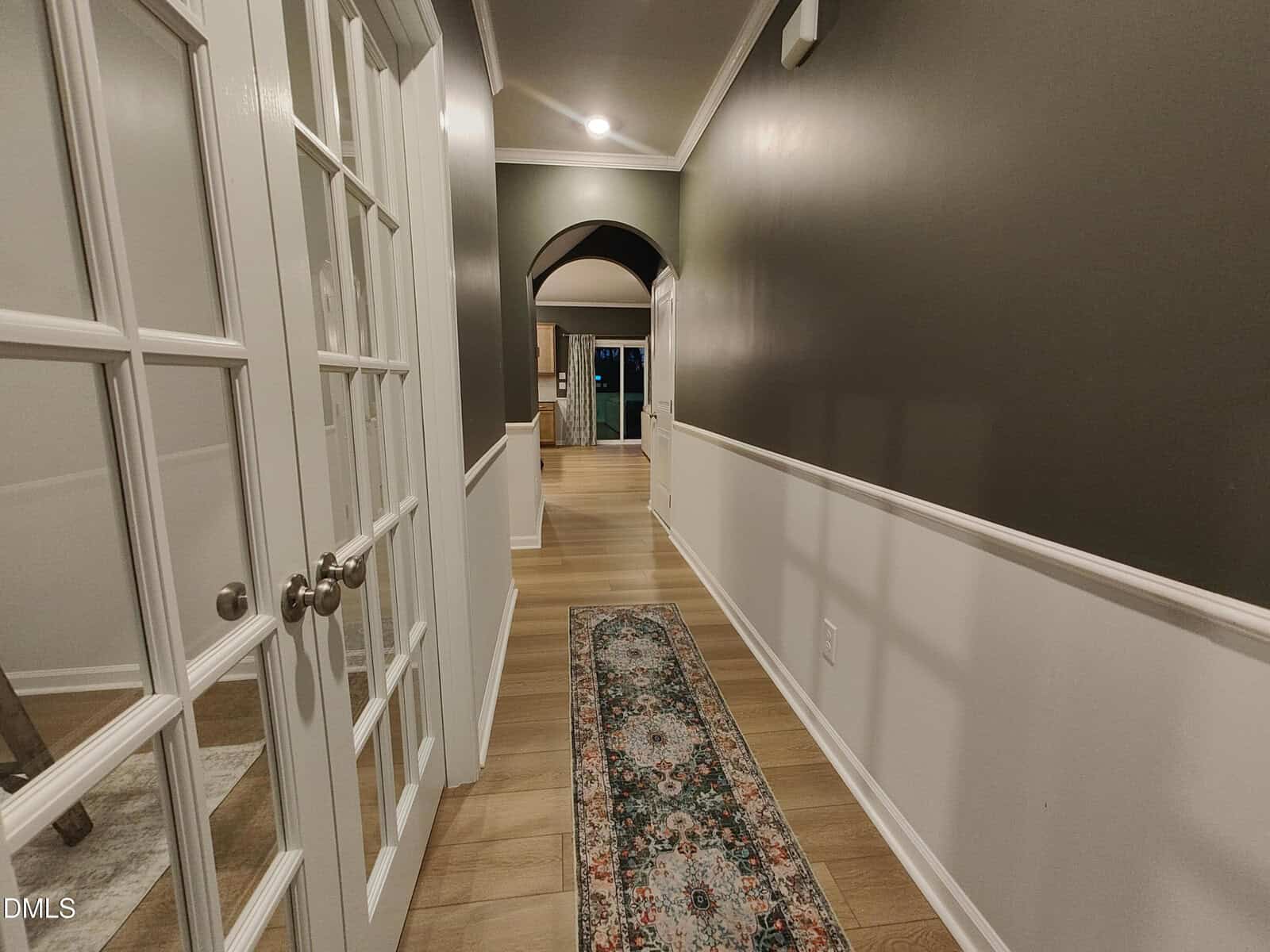 A hallway with wood flooring, a floral runner rug, dark upper walls, white lower paneling, and French doors on the left. An arched doorway leads to a well-lit room in the distance.