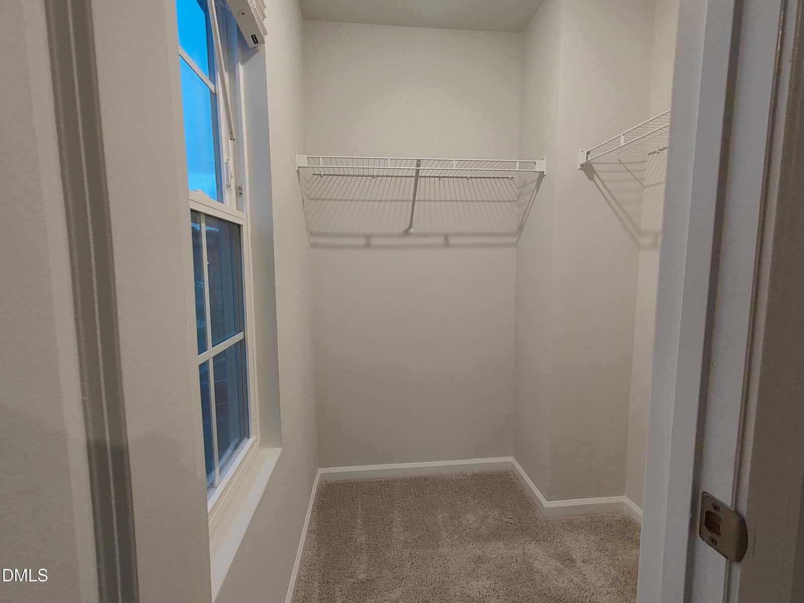 A small walk-in closet with beige carpet, white walls, a window on the left, and two white wire shelves on the right and back walls.