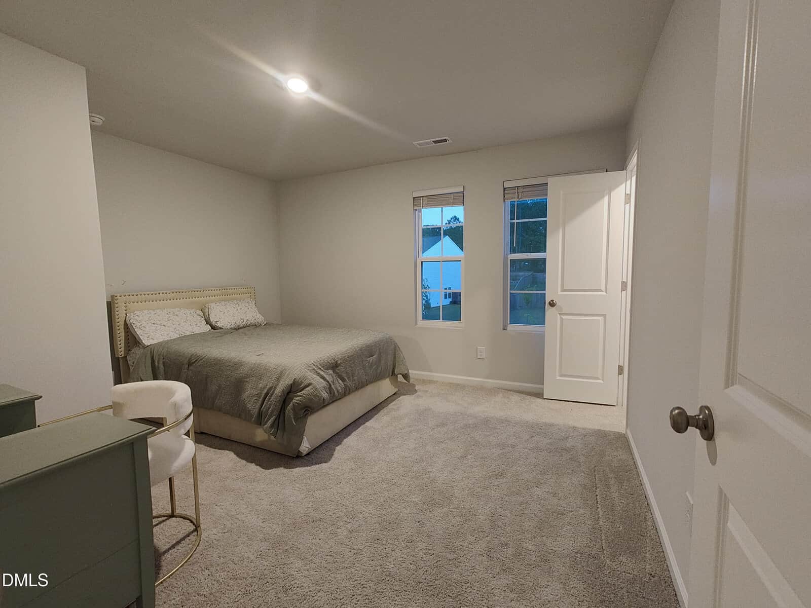 A simple bedroom with a bed, green dresser, and white chair. The room has gray carpet, light walls, two windows, and a door. Ceiling light is on, and houses are visible outside the windows.