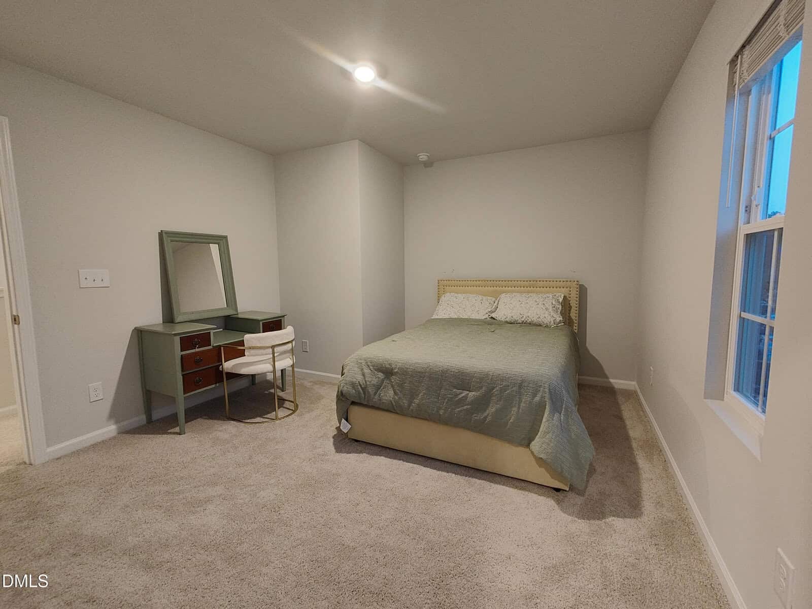A bedroom with beige carpet, a bed with green bedding, a green desk and chair with a mirror, white walls, and a window on the right letting in evening light.