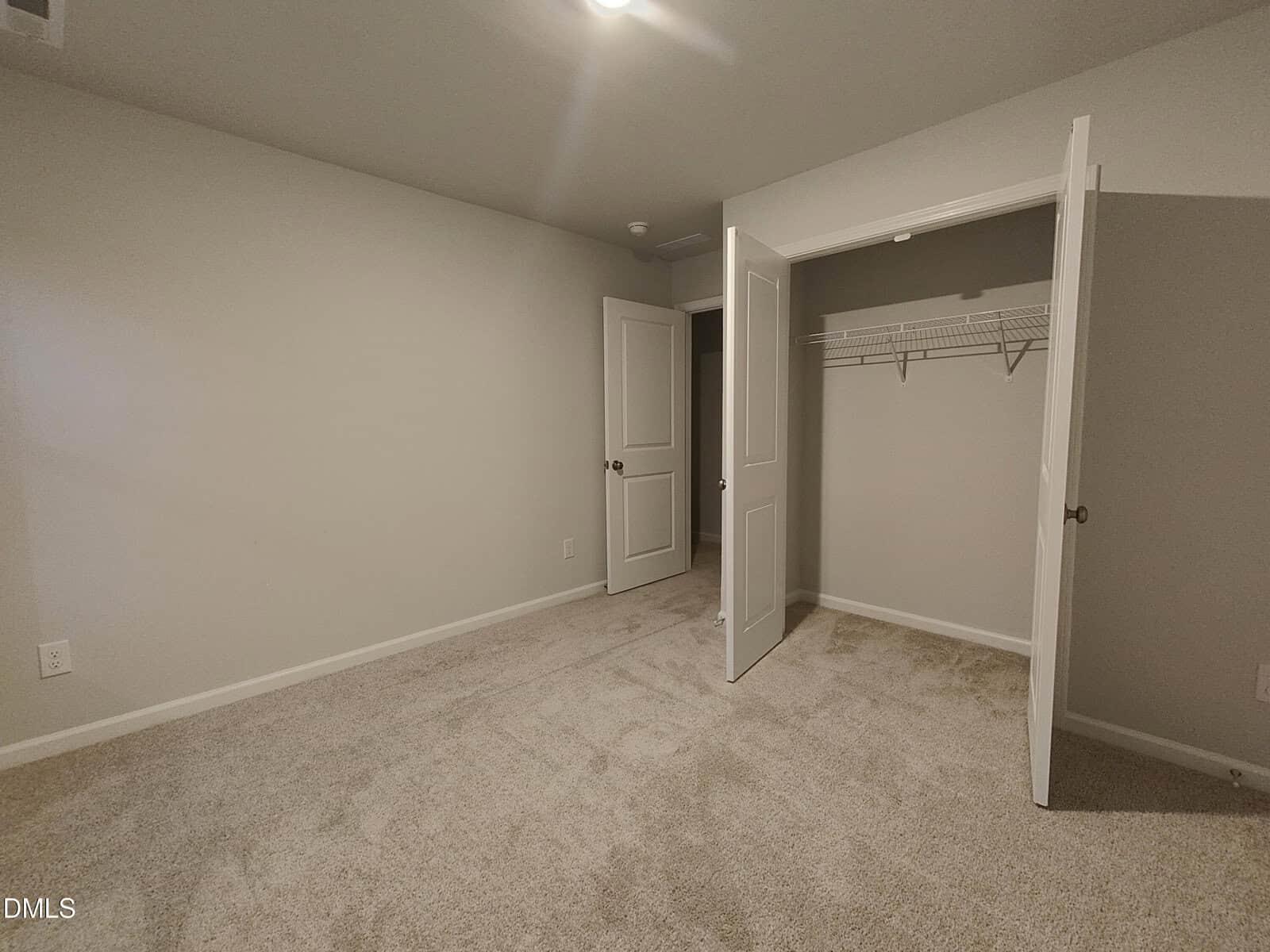 Empty bedroom with beige carpet, light gray walls, an open closet with a wire shelf, and an open door leading to another room. Ceiling light is on, and there are no windows visible.