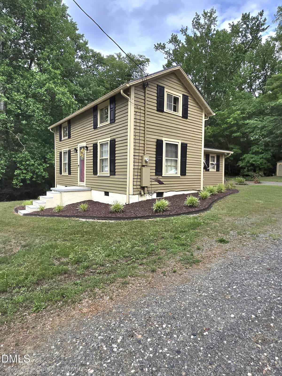 A two-story tan house with black shutters and a red door, surrounded by a small garden, sits next to a gravel driveway and is bordered by trees and grass.