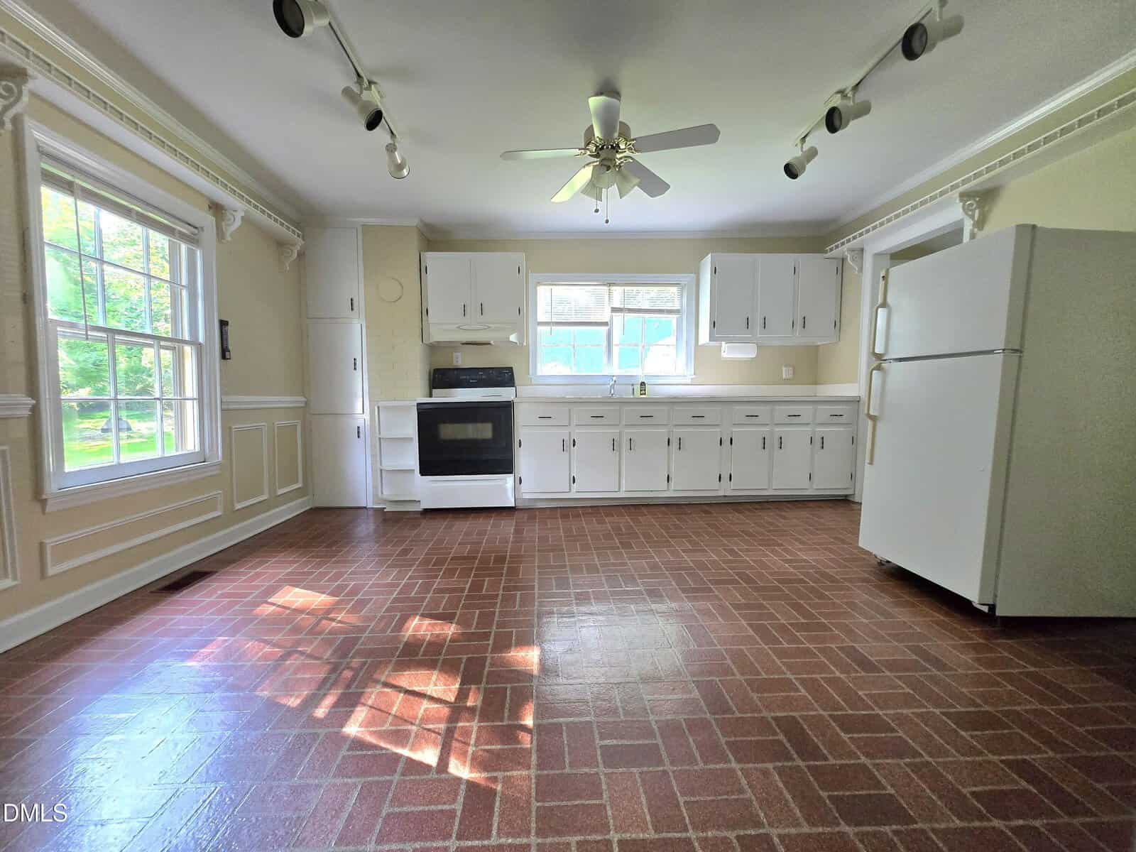 Bright kitchen with white cabinets, a ceiling fan, white refrigerator, oven, and a large window letting in natural light. Red brick-patterned floor and beige walls with decorative trim.