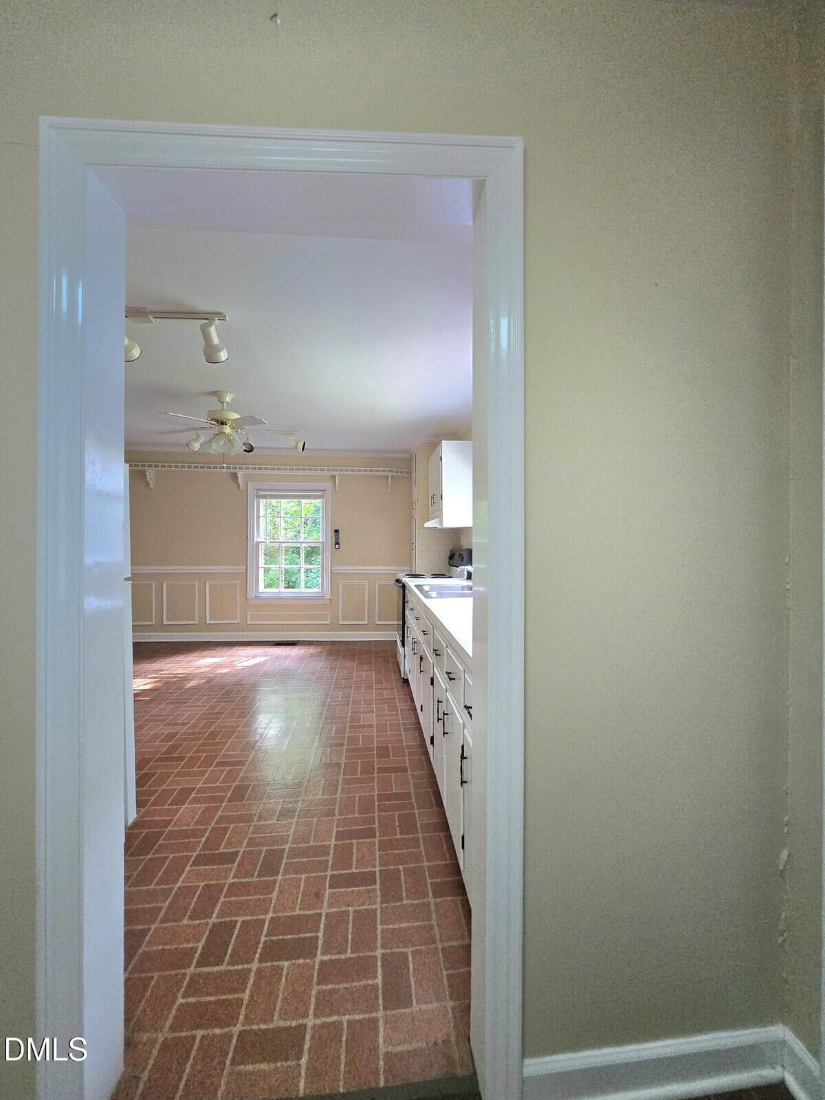 View through a doorway into a kitchen with brick-patterned flooring, white cabinets, and a window letting in natural light. The far room has beige walls, molding, and a ceiling fan with lights.