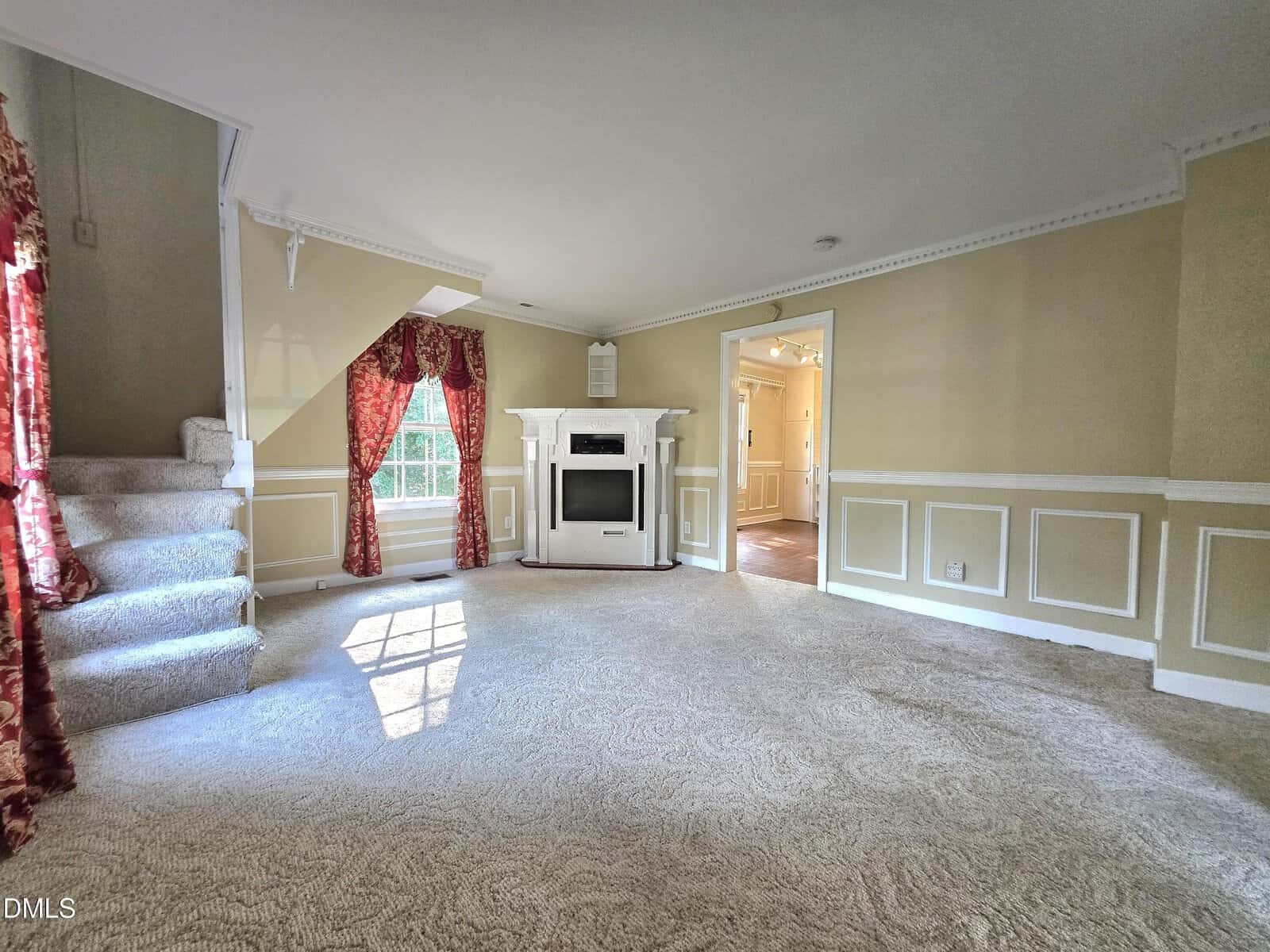 A bright, carpeted living room with cream walls, a white fireplace, red curtains, and a staircase on the left. A large window lets in natural light and there's an open doorway to another room.