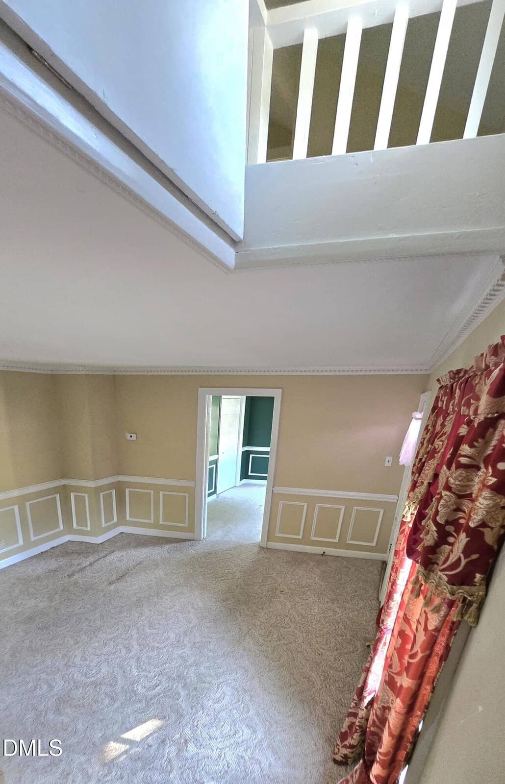 A view from a loft shows a carpeted room with beige walls, crown molding, red patterned curtains, and an open doorway leading to another brightly lit room.
