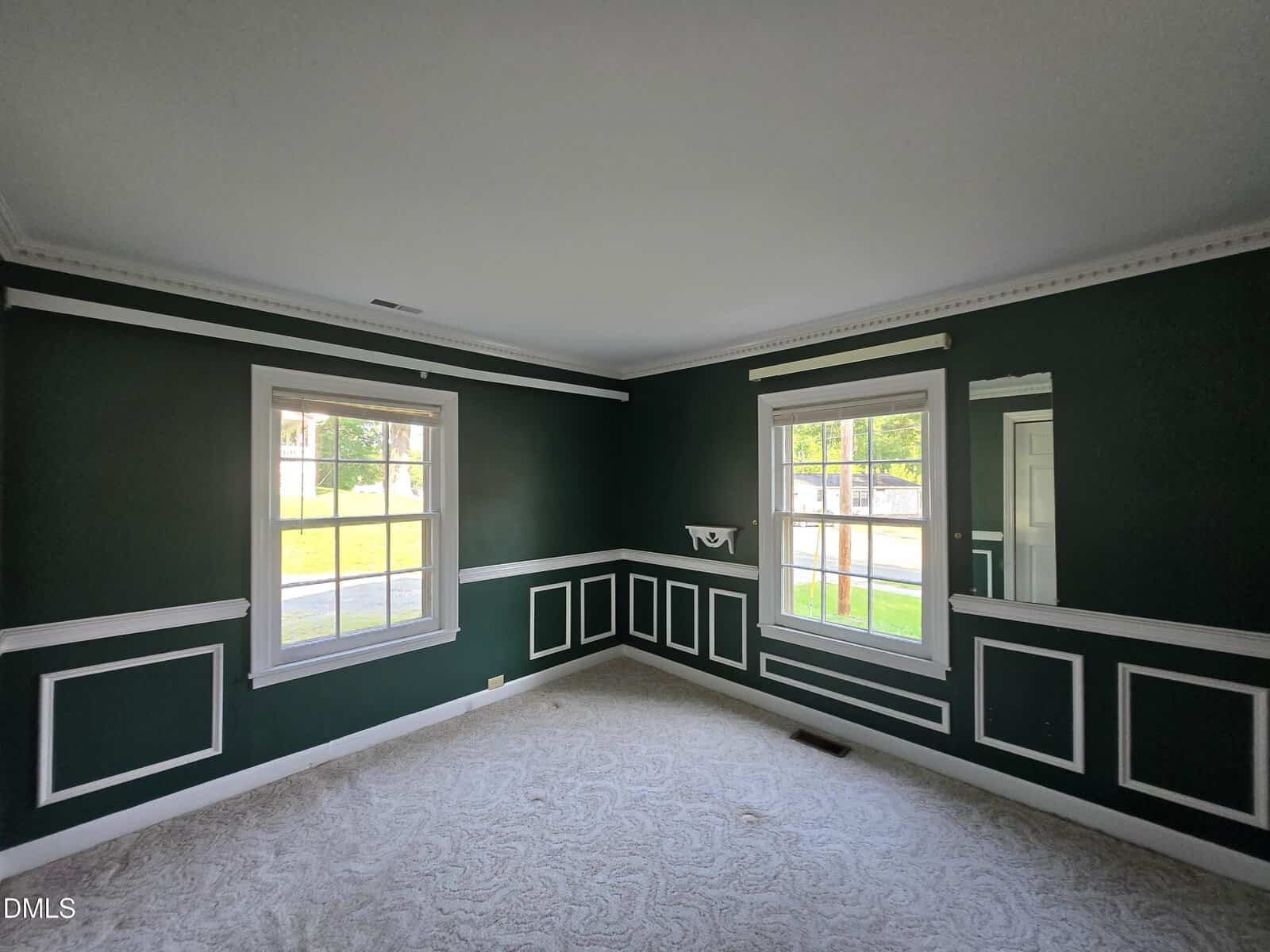 Empty room with dark green walls, white decorative trim, two large windows letting in sunlight, a small mirror, and light carpet flooring. The room appears clean and has detailed crown molding along the ceiling.