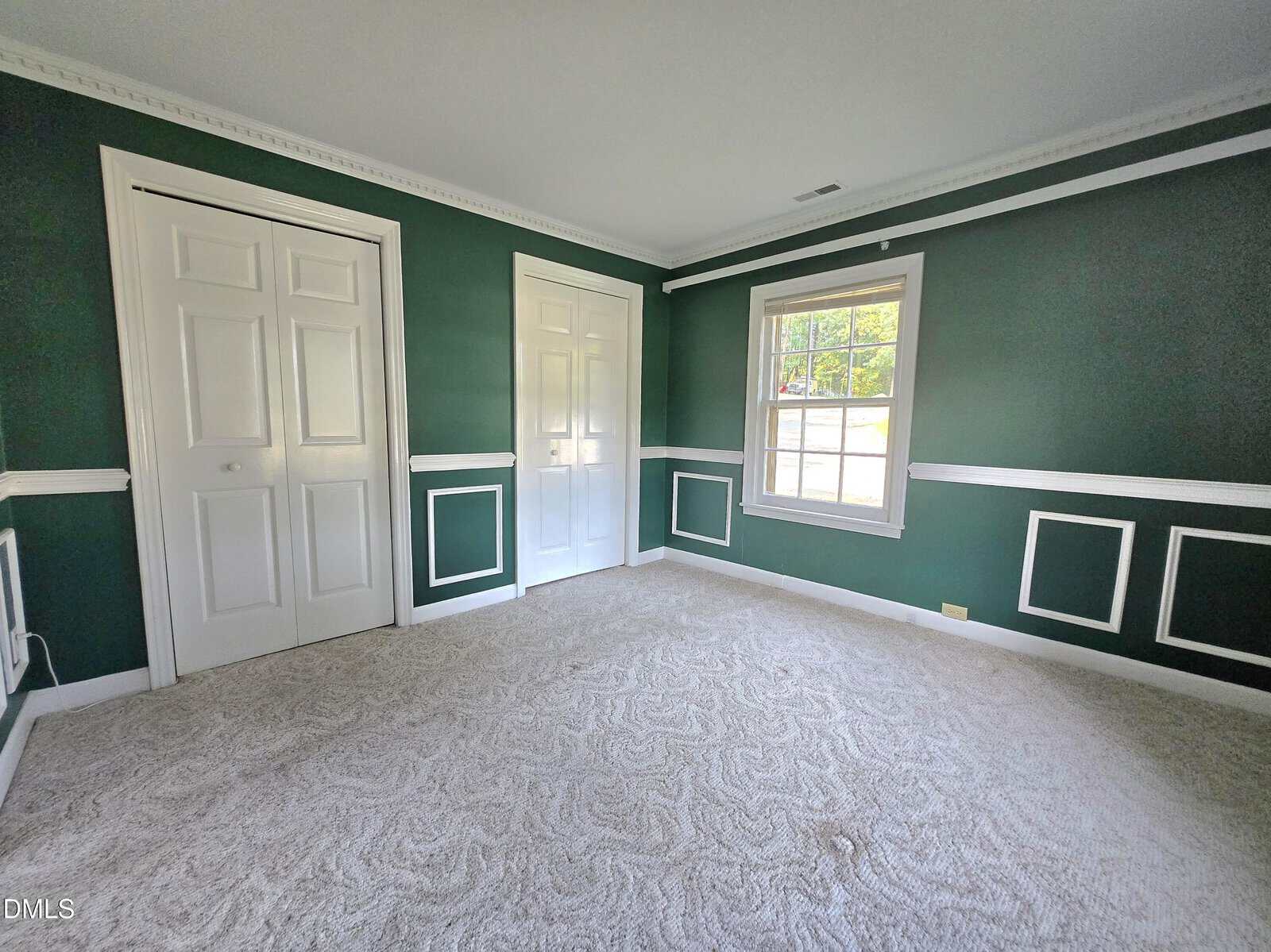 Empty room with dark green walls, white trim, two white closet doors, a large window letting in natural light, and light-colored carpet. Crown molding and wall frames add decorative detail.