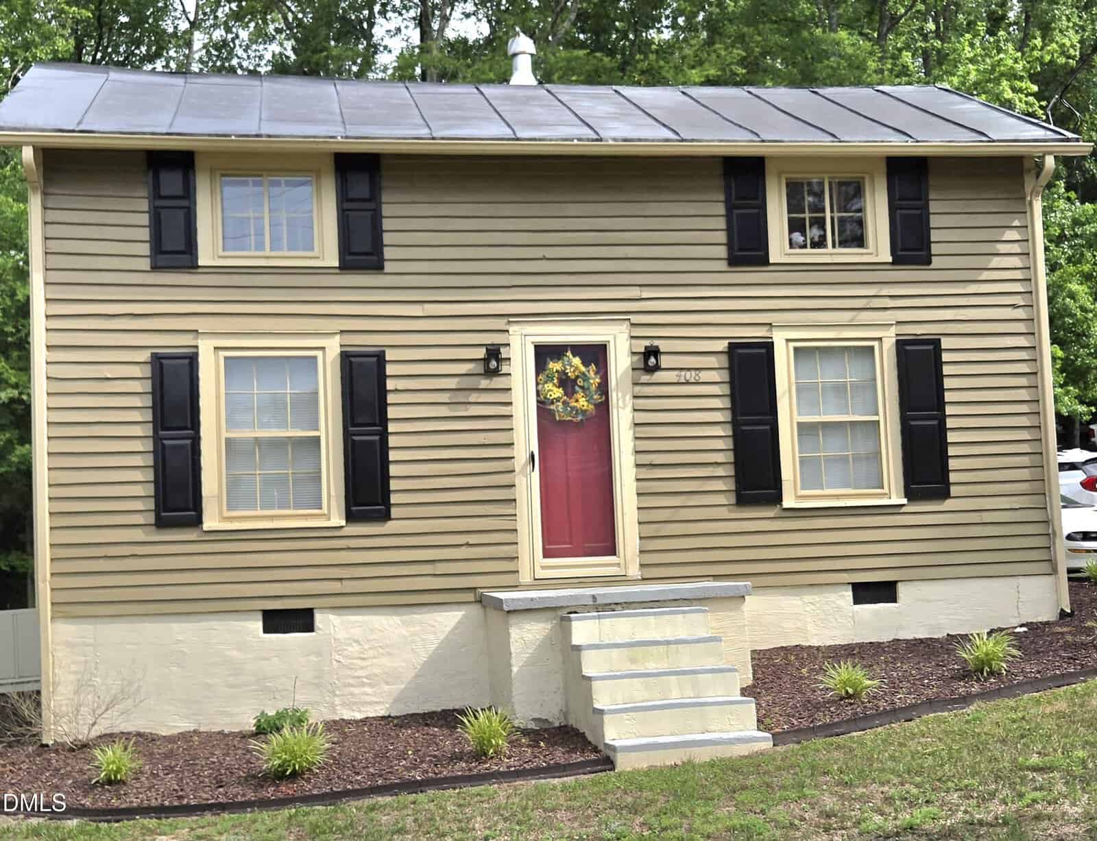 A small tan house with black shutters, a red front door decorated with a wreath, and a short set of white steps leading up sits at 408 N Durham Avenue in Creedmoor. The yard is landscaped with mulch and small green plants, while trees are visible in the background.