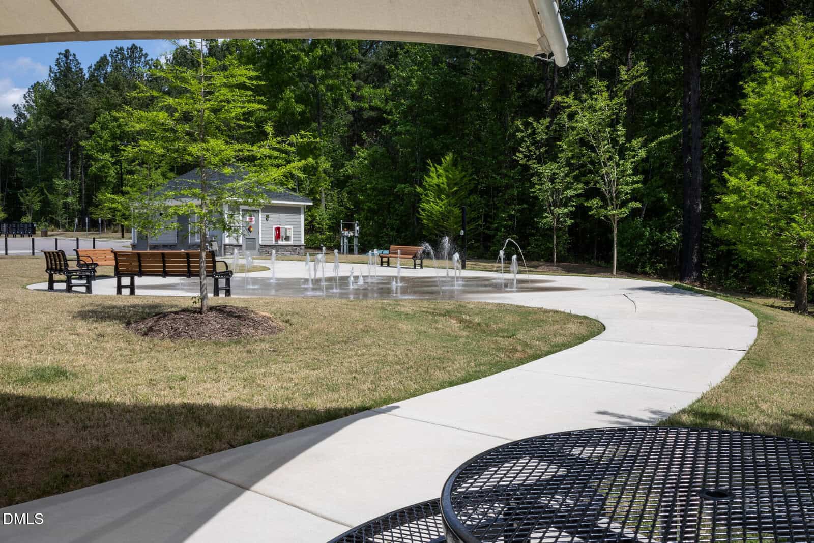 A small splash pad with water jets is surrounded by benches and a sidewalk. Trees and a small building are in the background. A metal table and chair are in the foreground under a shaded canopy.