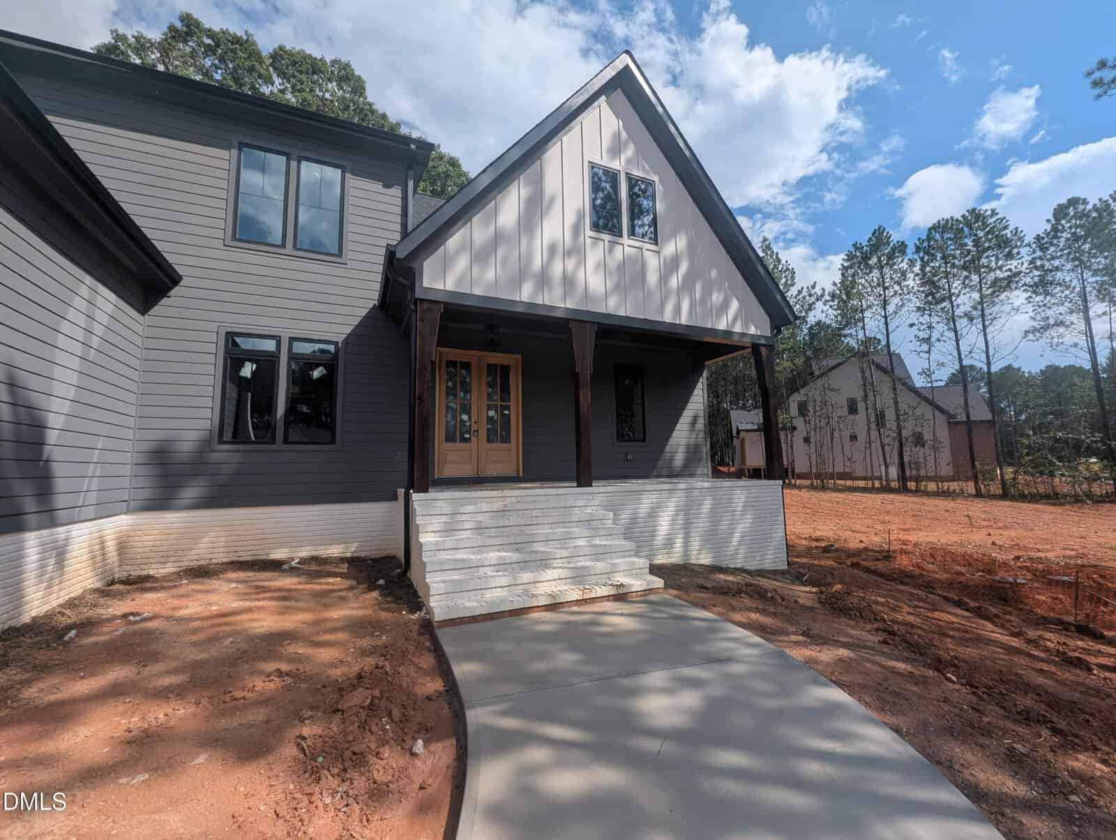 Modern gray house with a covered front porch and wooden double doors. Concrete walkway leads to steps at the entrance. Yard is unfinished with exposed soil, and another house is visible in the background among trees.