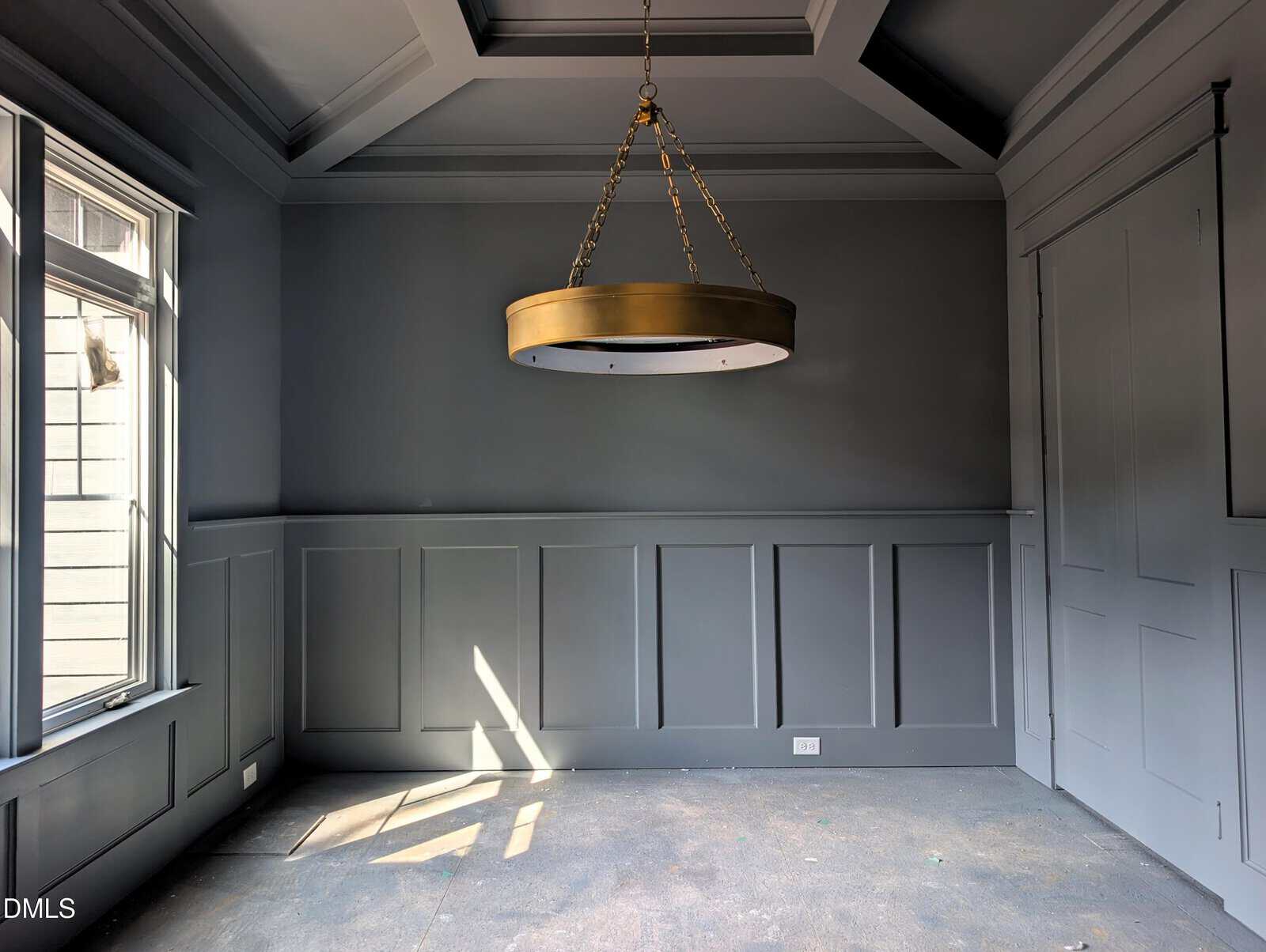 A gray, empty room with wainscoting, a large circular brass chandelier, a window letting in sunlight, and uncarpeted floors.