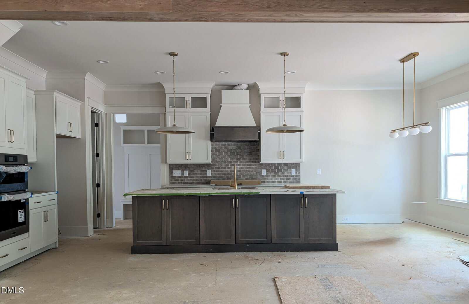 Modern kitchen under construction with white cabinets, a center island with dark wood base, two pendant lights above, tile backsplash, and large windows letting in natural light. Floor appears unfinished.