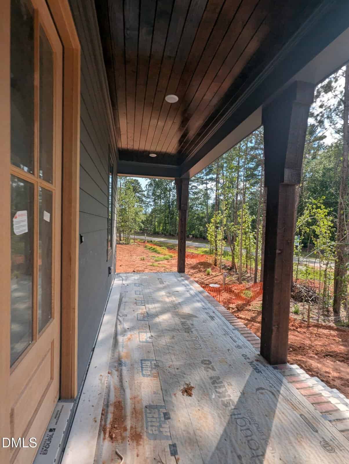 A covered front porch of a house under construction, with exposed building materials on the floor, wooden columns, dark ceiling, and view of dirt yard and trees in the background.