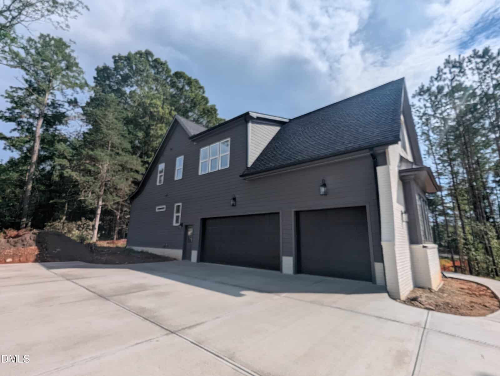 Gray two-story house with three garage doors, white trim, and black roof, surrounded by trees and set on a large concrete driveway under a partly cloudy sky.