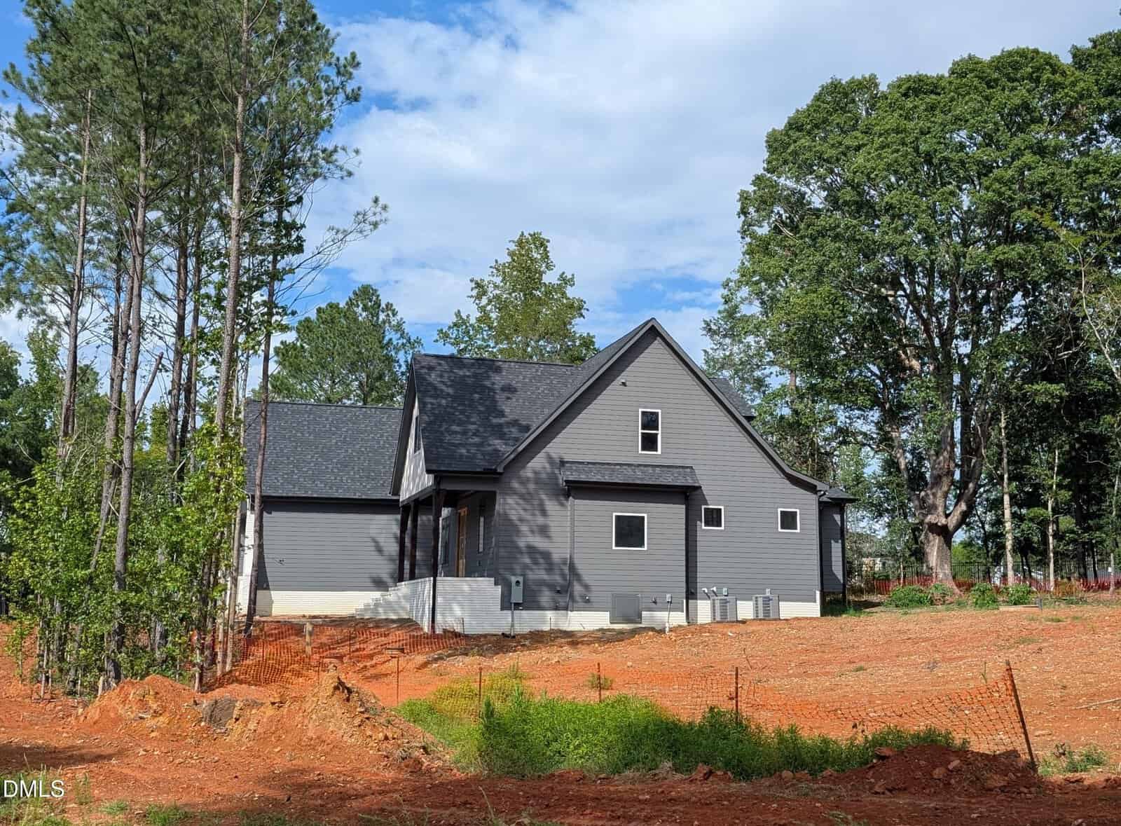 A modern gray house with black roof sits on a plot of red dirt, surrounded by tall trees and some green grass. The sky is partly cloudy and a small fence runs across the yard.