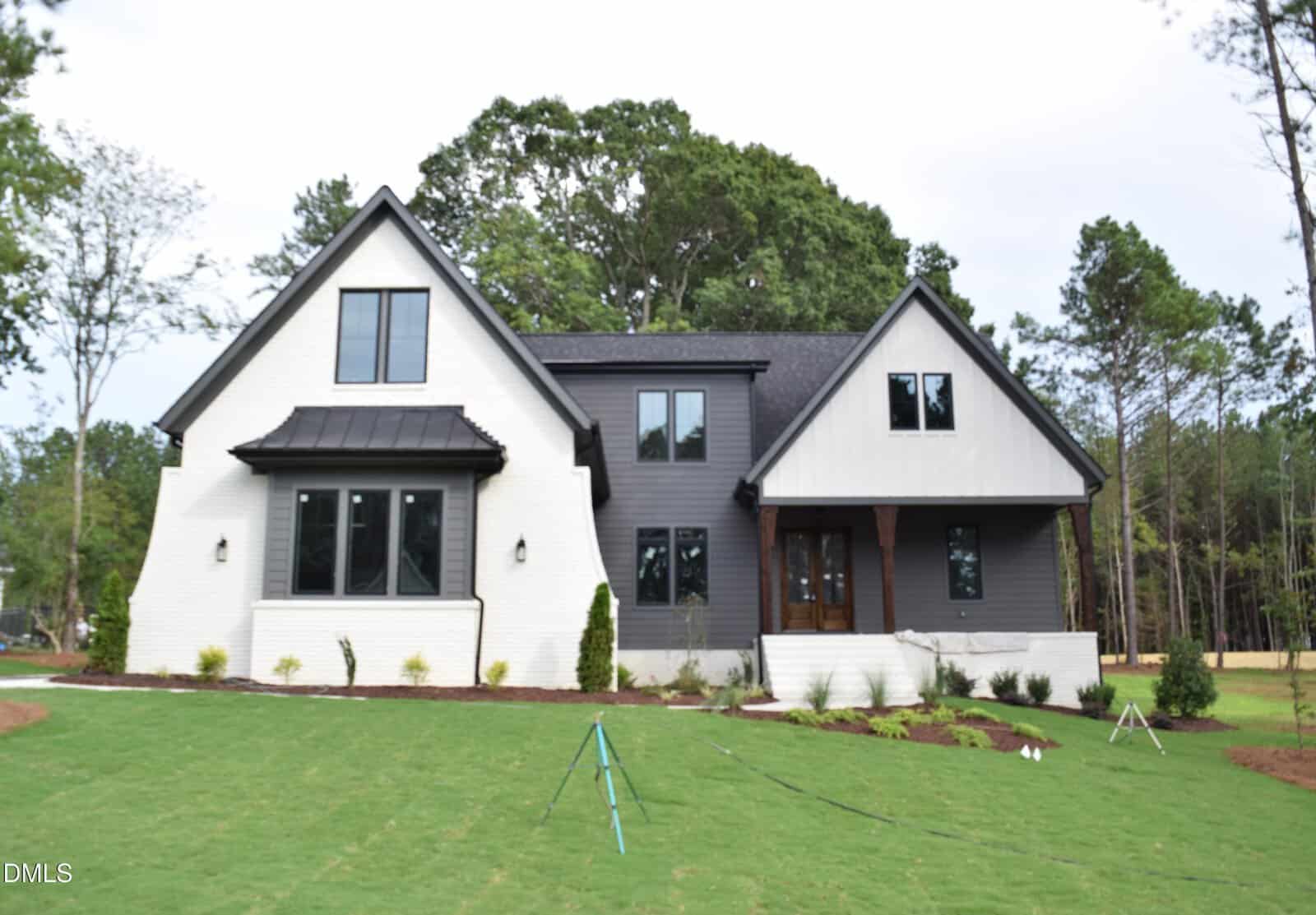 A modern two-story house at 1208 Red Cedar Court, Youngsville features white brick and dark gray siding, large windows, and a sloped roof. It sits on a freshly landscaped lawn with small shrubs and is surrounded by tall trees.