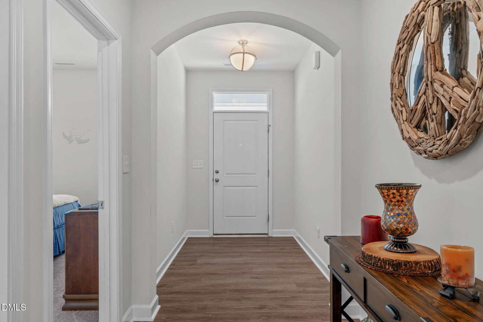 A hallway with wood flooring, a white front door, an arched entryway, a ceiling light, and a table with decorative items and a round mirror on the wall. A glimpse of a bedroom is visible on the left.