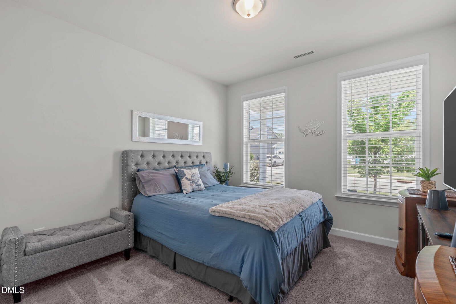 Bright bedroom with a blue bed, gray tufted bench, wall mirror, dresser with plants, and large windows letting in natural light. Walls and carpet are light neutral colors.