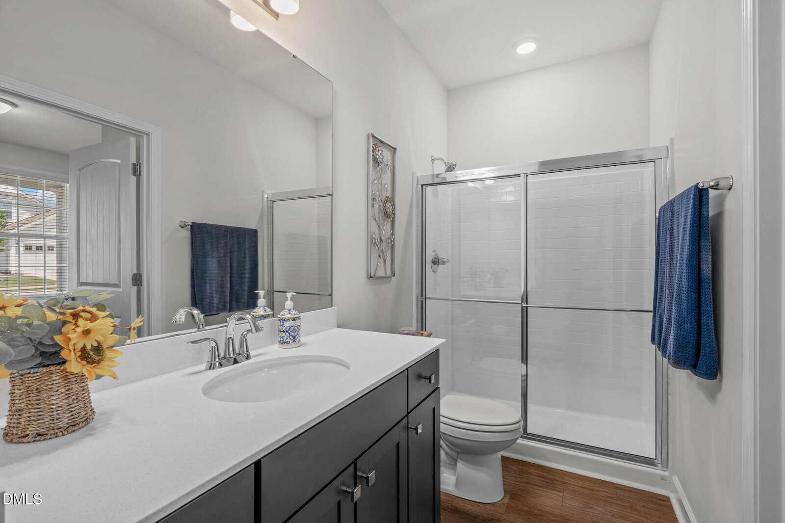 Modern bathroom with white countertop, dark cabinets, a sink, large mirror, toilet, and glass shower. Blue towels hang on both sides, and a vase with sunflowers decorates the counter.