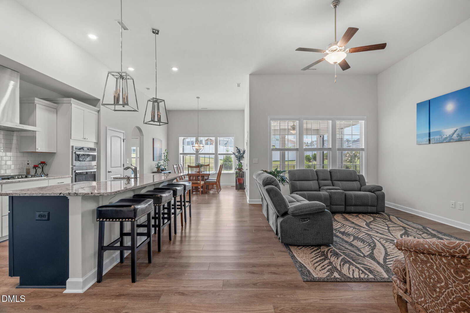 Open-concept living area with modern kitchen, barstools, dining table, gray sectional sofa, ceiling fan, and large windows letting in natural light. Light walls and wood floors create a bright, inviting space.