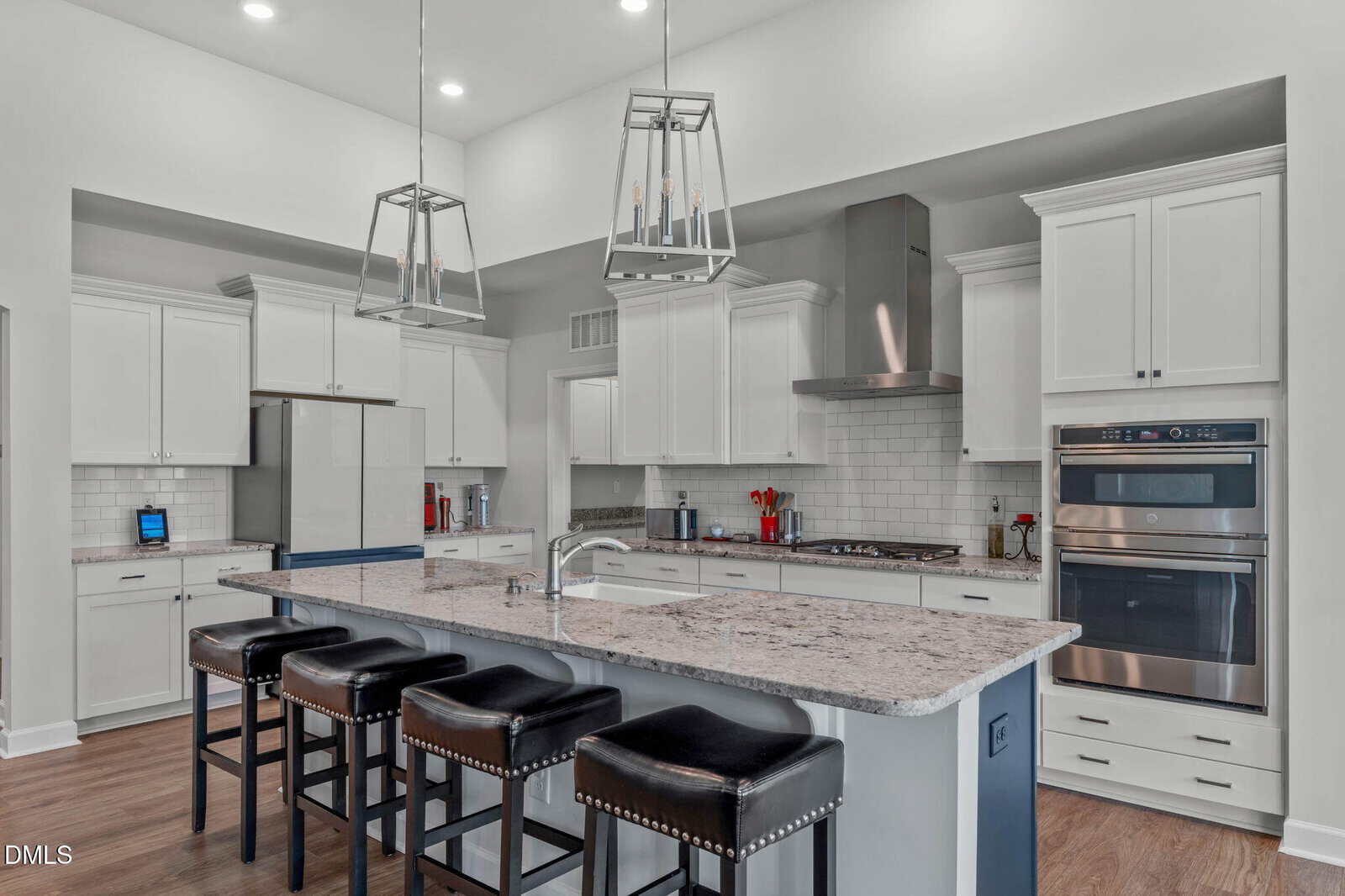 Modern kitchen with white cabinets, stainless steel appliances, two pendant lights, a large island with granite countertop, and four black leather stools on wooden flooring.
