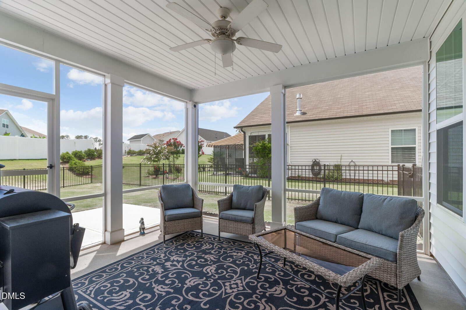 Screened porch with wicker furniture and a glass coffee table on a patterned rug, ceiling fan above. Green backyard with a fence, grill, and two neighboring houses visible outside.