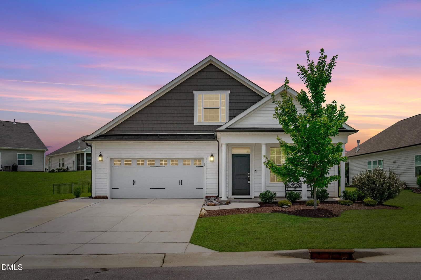 Single-story suburban house with a gray roof, white siding, and a two-car garage, set against a colorful sunset sky. Located at 45 Moon Flower Walk in Youngsville, the property features a young tree and neatly trimmed bushes in the front yard.