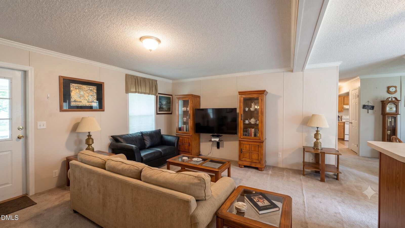 A cozy living room at 4552 Gray Rock Road, Kittrell, with two sofas, a coffee table, TV on a wooden stand, two lamps, and framed art. The space leads into a kitchen and features beige walls and carpeting.