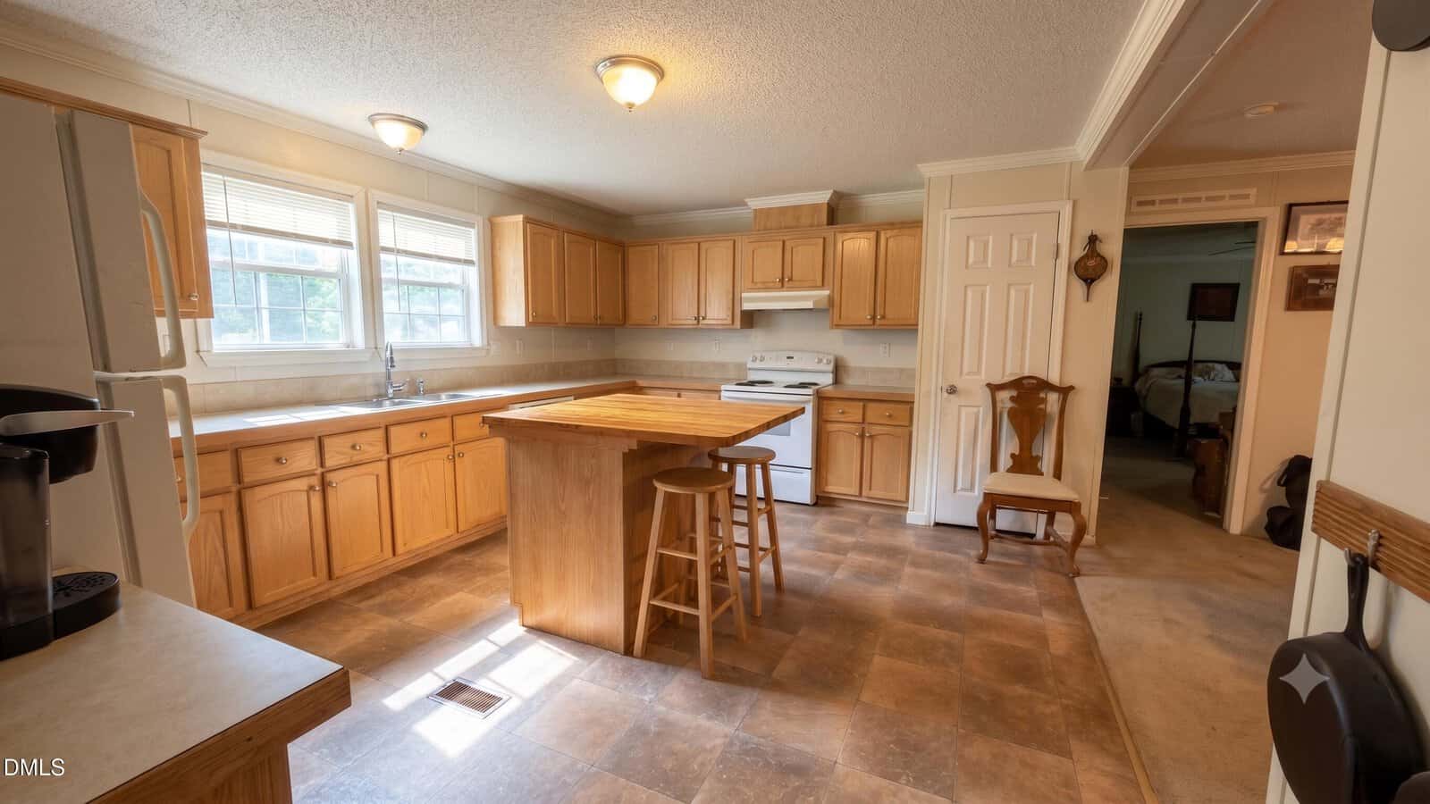 Spacious kitchen with wooden cabinets, a central island with two stools, white appliances, and sunlight streaming through three large windows. Adjacent room with a chair is visible through an open doorway.