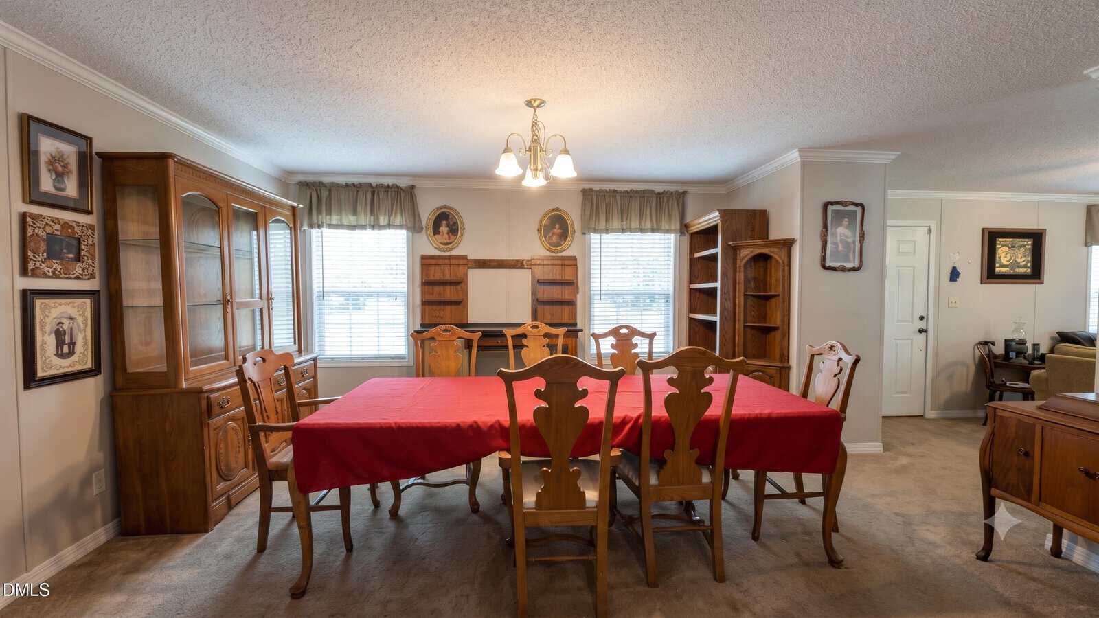 Dining room with a large wooden table covered by a red tablecloth, six chairs, wooden cabinets, shelves, framed art on the walls, and two windows with curtains letting in natural light.