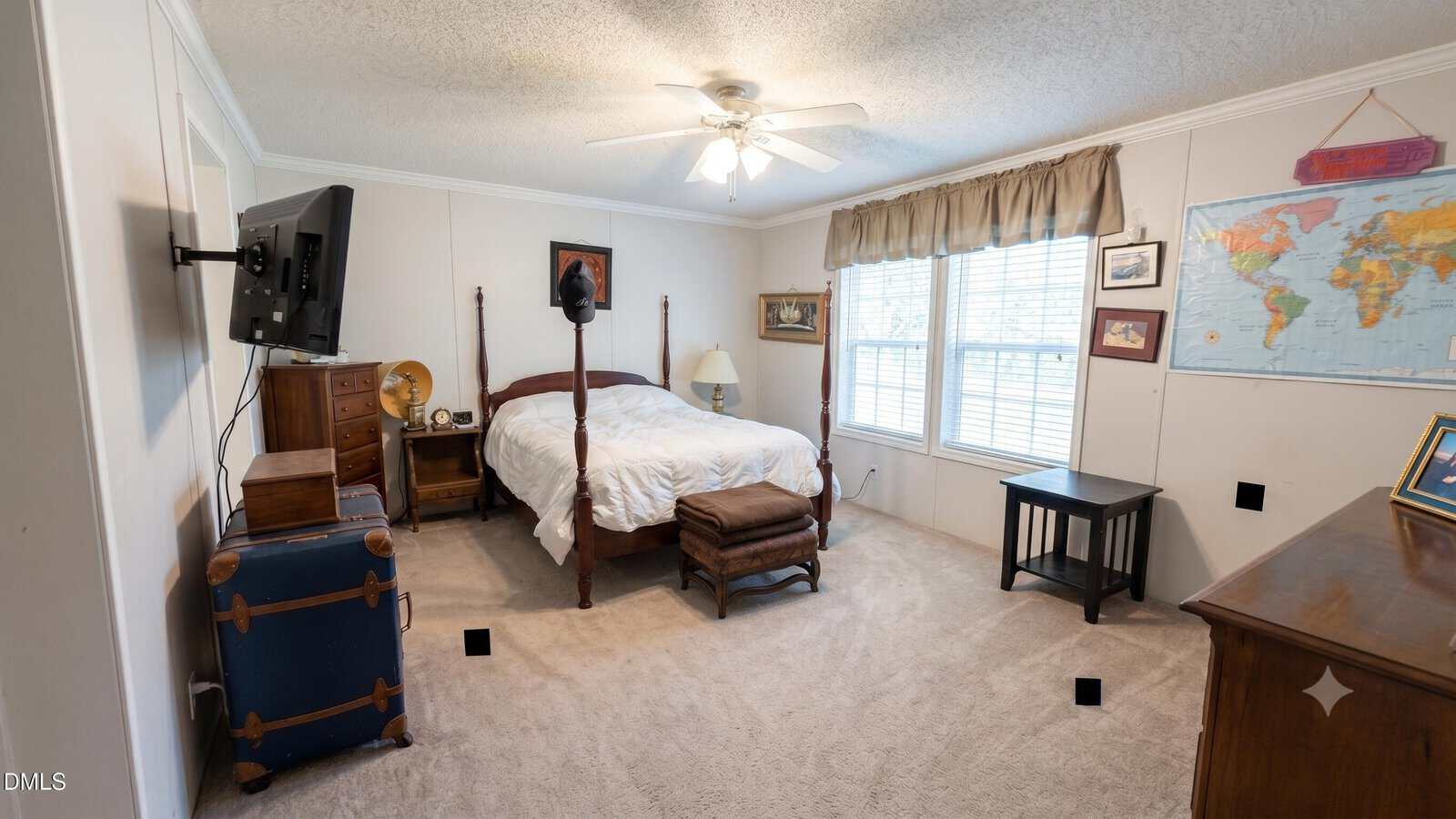 A bedroom with a four-poster bed, wooden furniture, a TV mounted on the wall, a blue trunk, a map on the wall, and large windows with blinds letting in natural light.