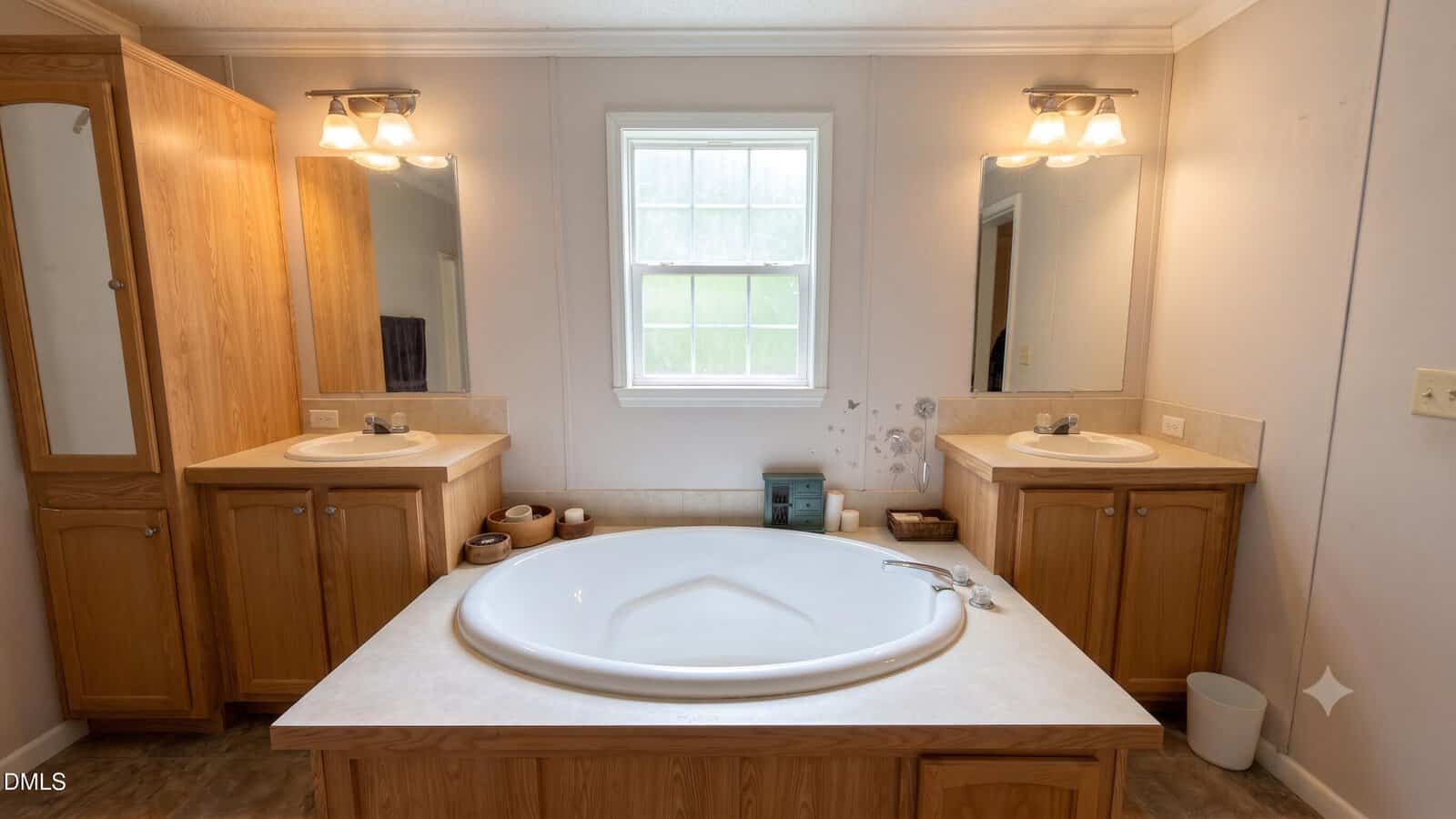 A bathroom with a large round bathtub centered between two wooden vanities, each with a sink, mirror, and light fixture. A window is above the tub, and various toiletries are on the counters.