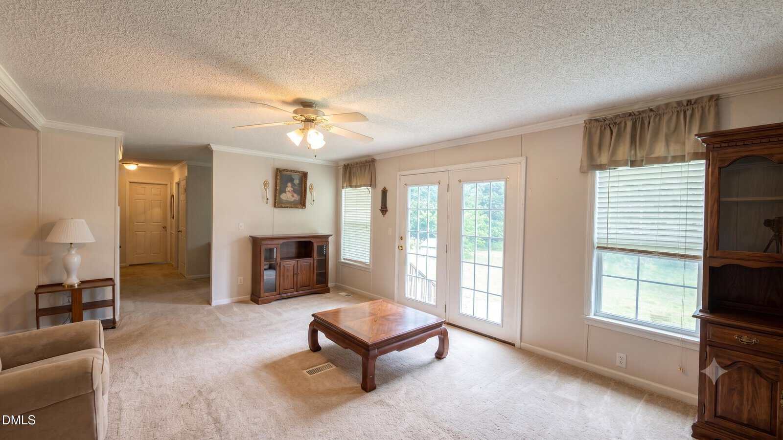 Carpeted living room with a ceiling fan, coffee table, glass doors, two large windows with curtains, a wooden cabinet, a side table with a lamp, and a hallway in the background. Natural light fills the room.