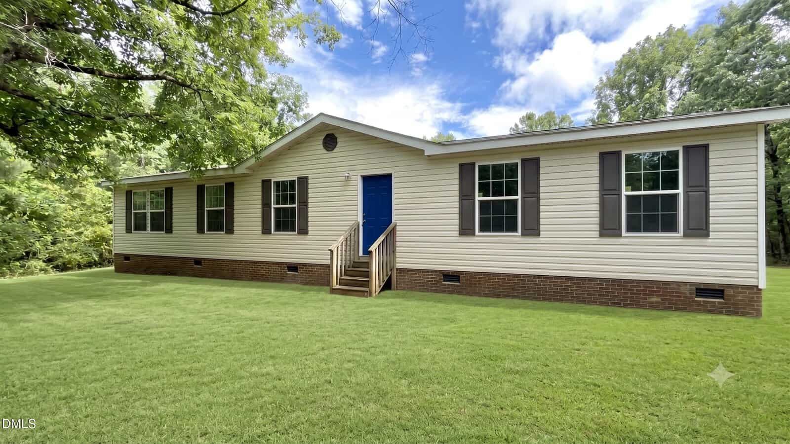 Charming single-story beige house with black shutters, a blue front door, and a small porch with steps at 4552 Gray Rock Road. This Kittrell home is surrounded by green grass, mature trees, and set beneath a partly cloudy sky.