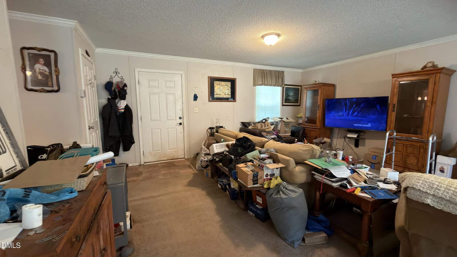 A cluttered living room with boxes, papers, and various items scattered on tables and the floor. A TV is mounted on the right wall, and a couch faces it. The front door and a window are at the back of the room.